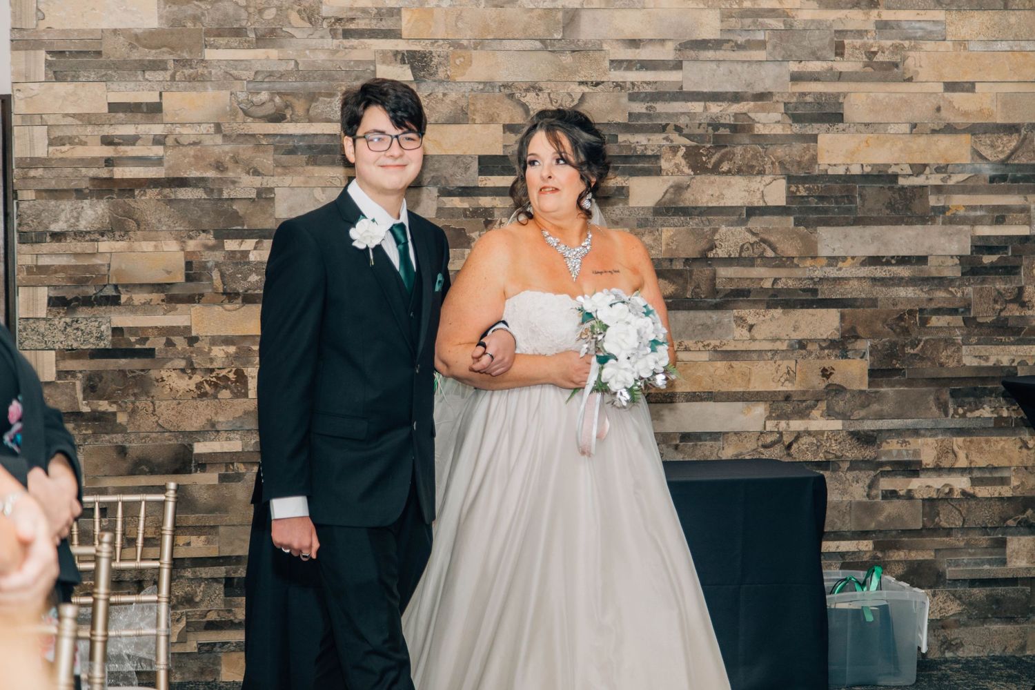 Bride in white gown holding bouquet walks down aisle escorted by young groomsman at wedding ceremony.