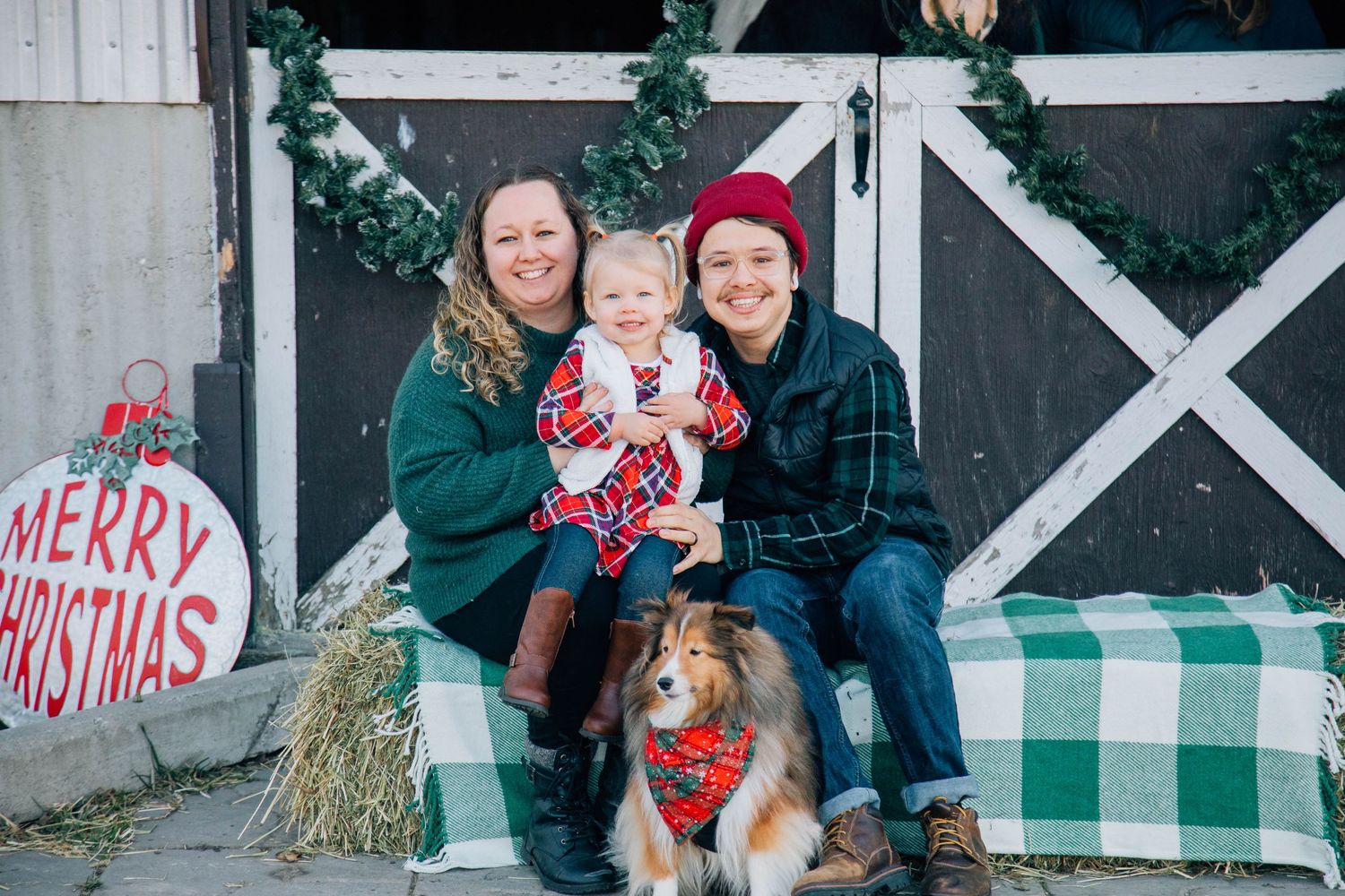 Family Christmas photo with two women, toddler, and Sheltie dog posed in front of decorated barn.