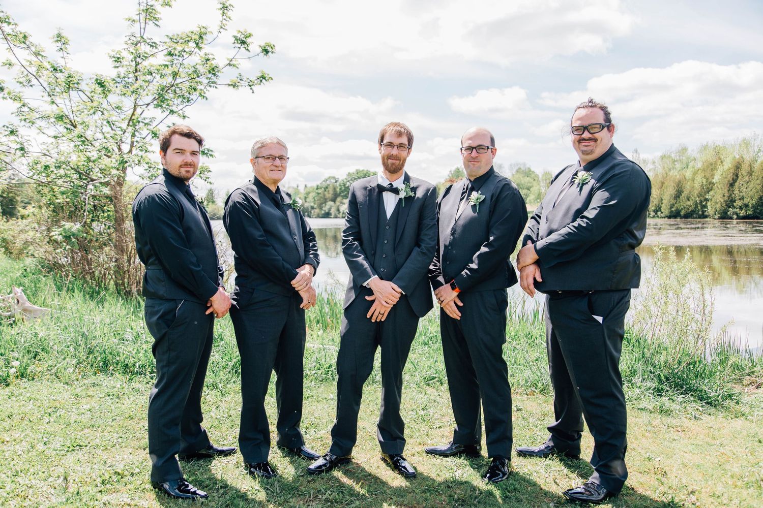 Five groomsmen in black tuxedos posing outdoors near a pond with green trees in background.