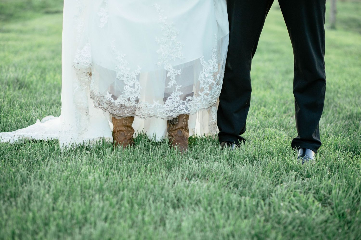 Close-up of bride wearing cowboy boots under her lace wedding gown on green grass.