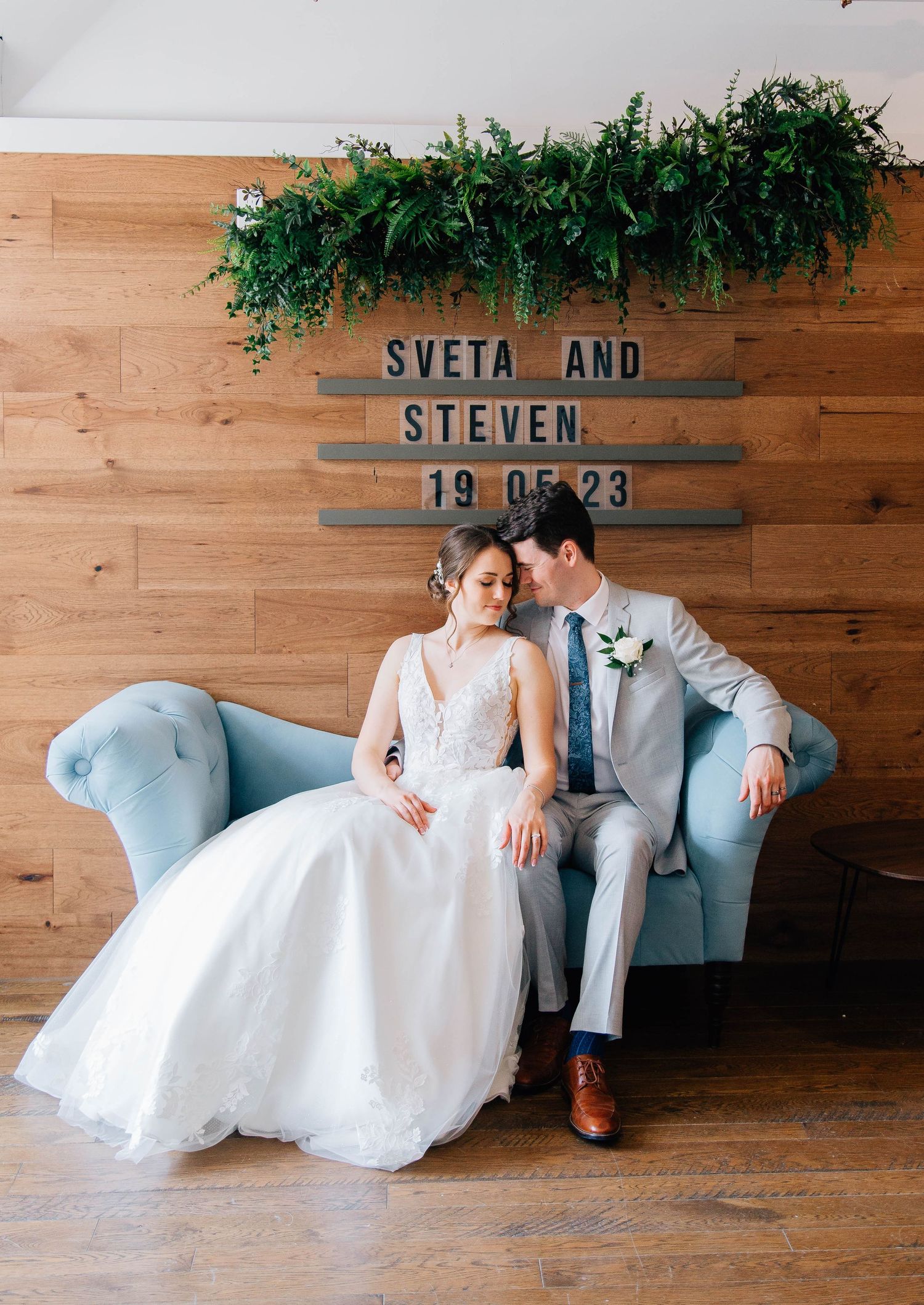 Bride and groom sitting together on a blue velvet sofa beneath a Sveta and Steven wedding date sign.