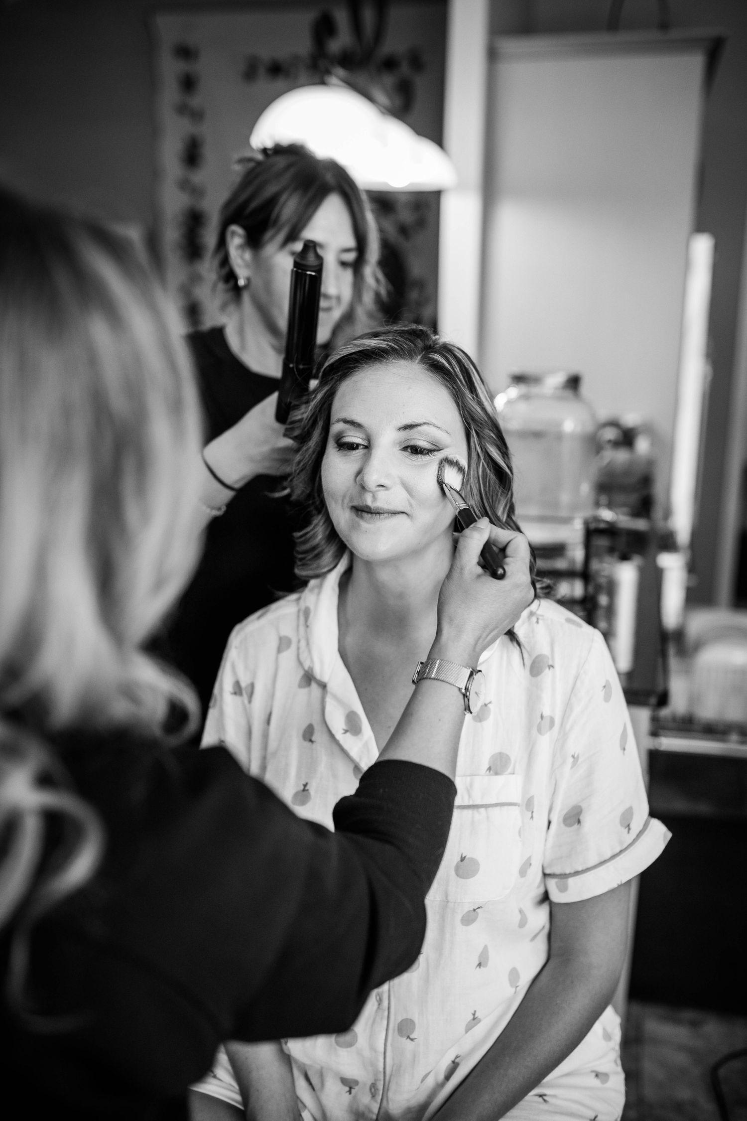 Black and white photo of a bride getting hair and makeup done by two stylists before her wedding ceremony.