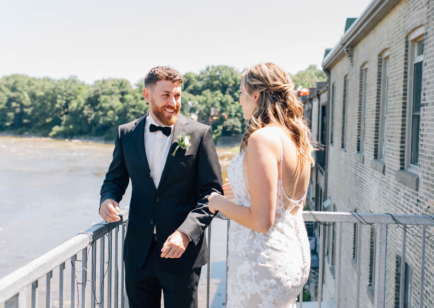 Groom in black tuxedo smiling at bride during sunny outdoor first look on balcony.