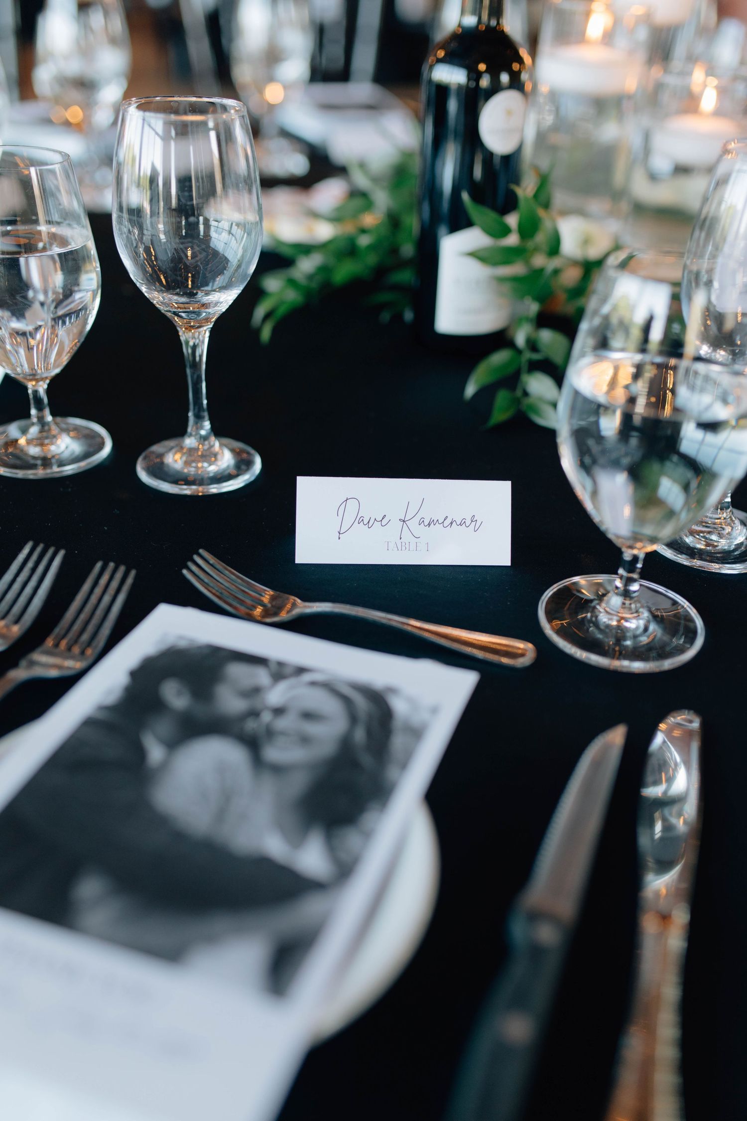Elegant wedding place setting with name card, black-and-white couple photo, wine glasses, and gold cutlery on dark tablecloth.