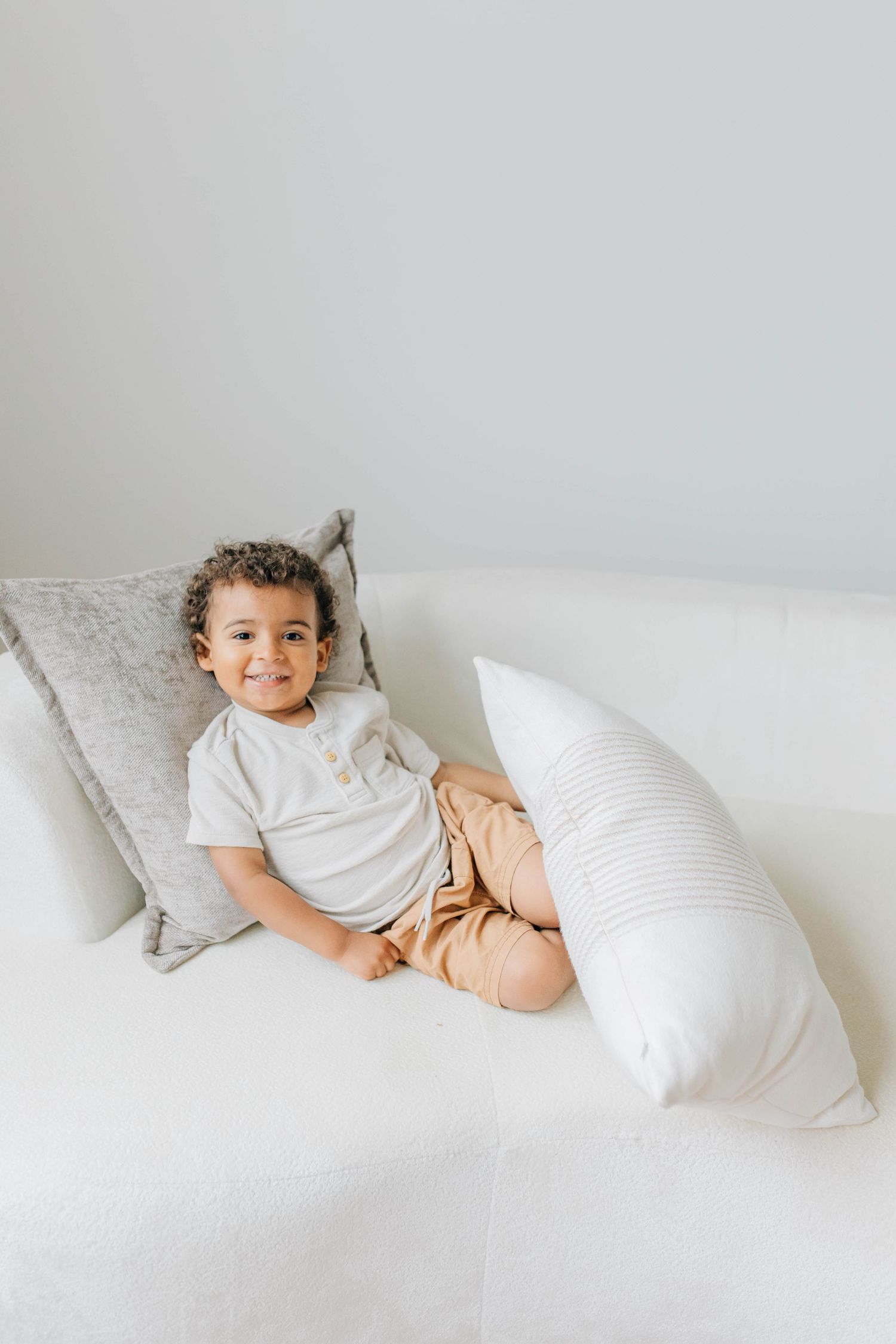 Adorable curly-haired toddler boy smiling while relaxing on white couch with pillows.