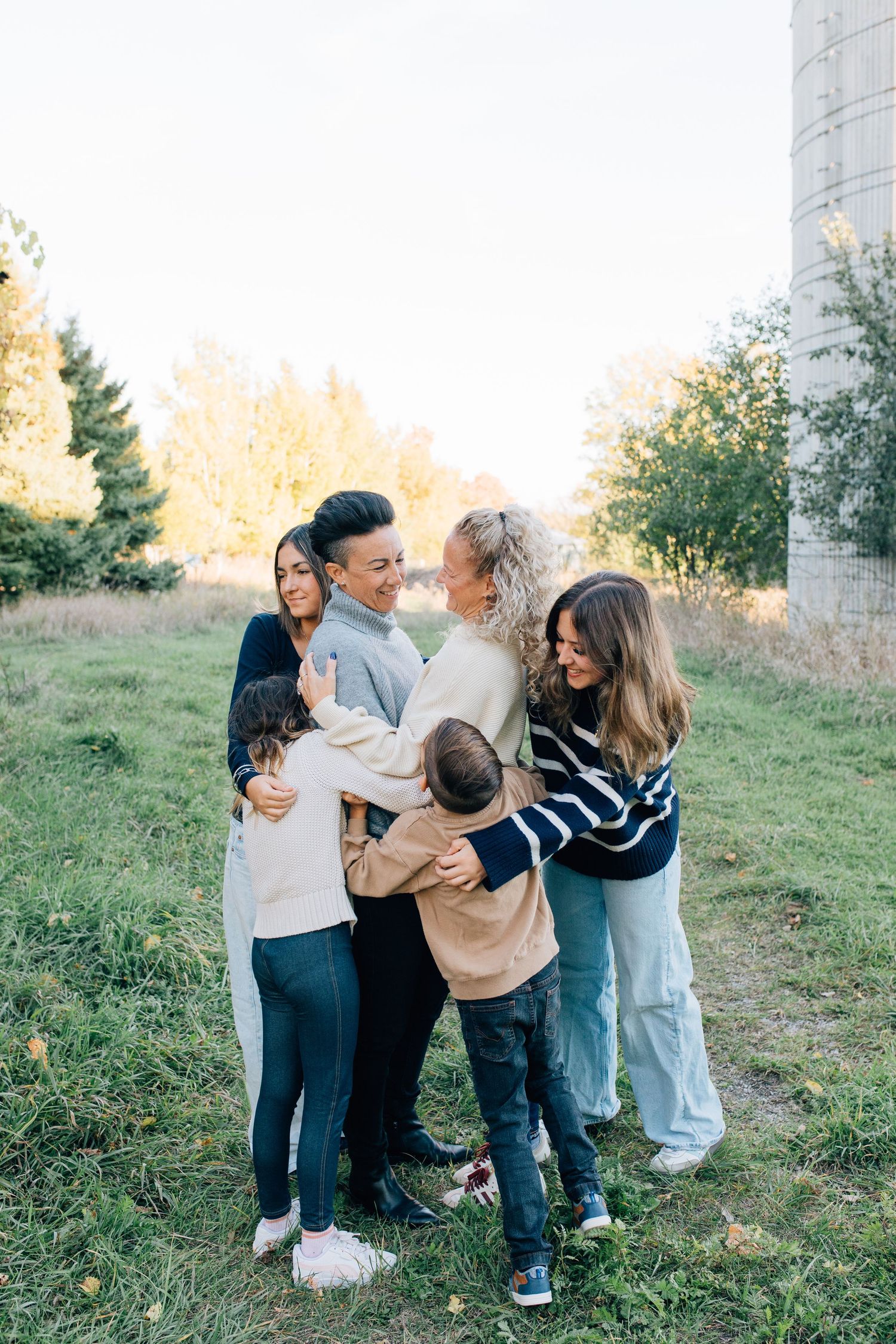 A joyful multigenerational family group hugging together outdoors in a green field during golden hour.