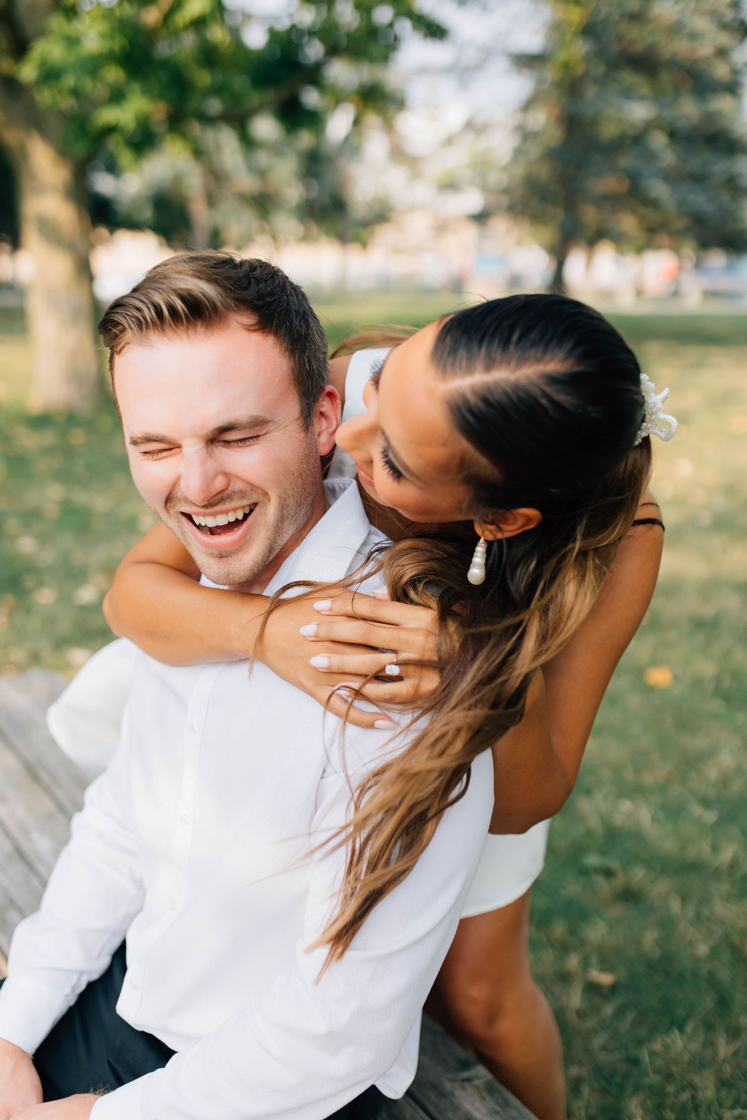 Joyful couple laughing outdoors, woman in white dress hugging man from behind in sunny park setting.