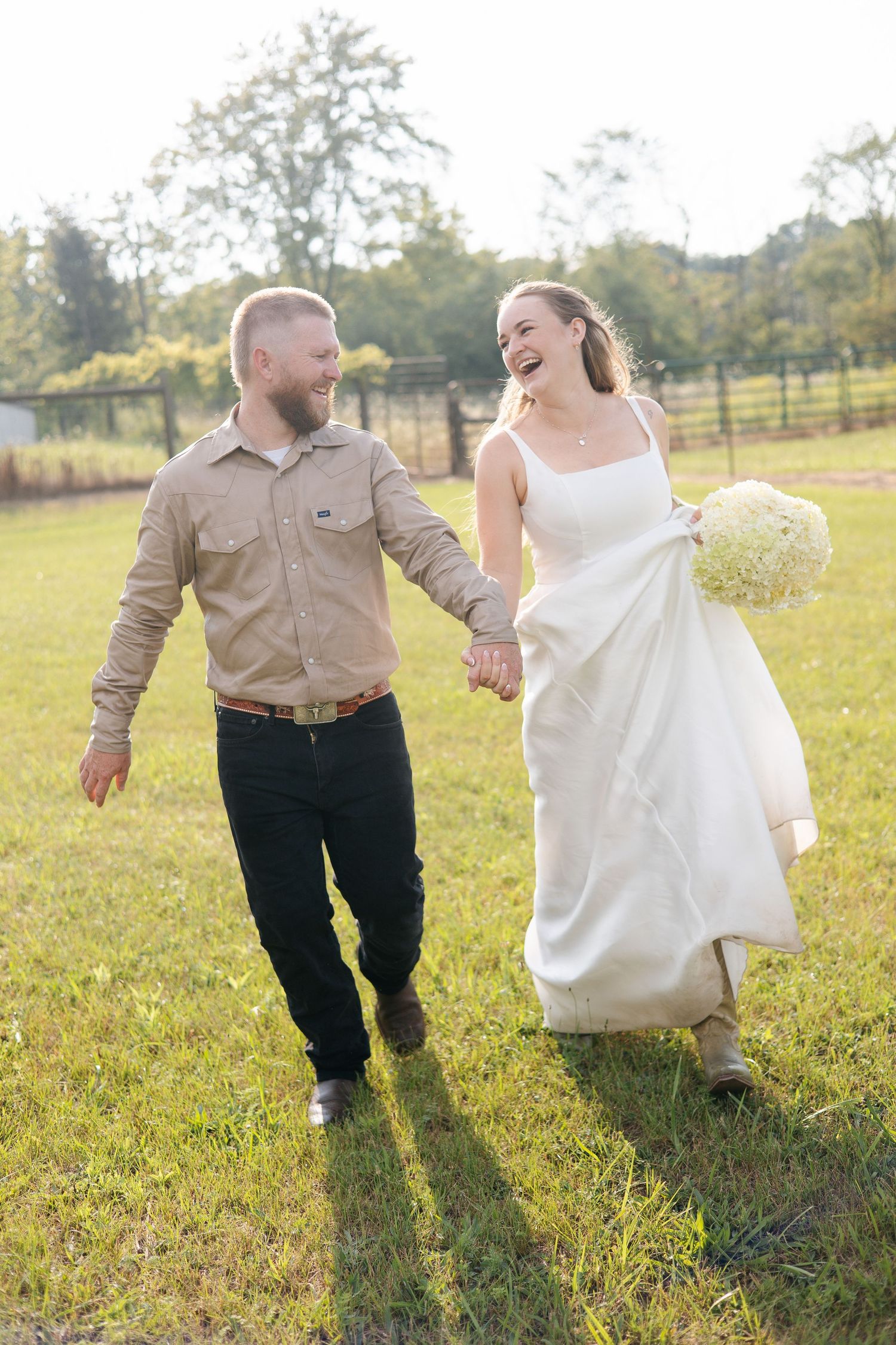 Happy bride and groom walking hand-in-hand through green field, bride holding white bouquet in wedding dress.