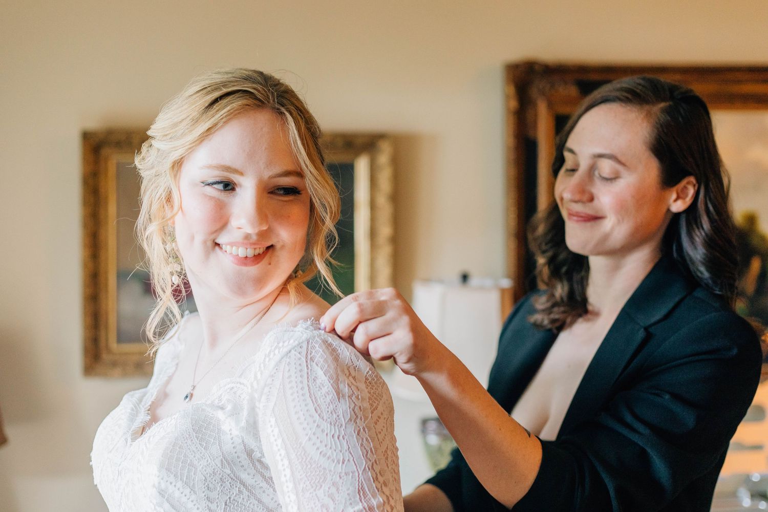 A bride smiles warmly as her partner helps fasten the back of her lace wedding dress before the ceremony.