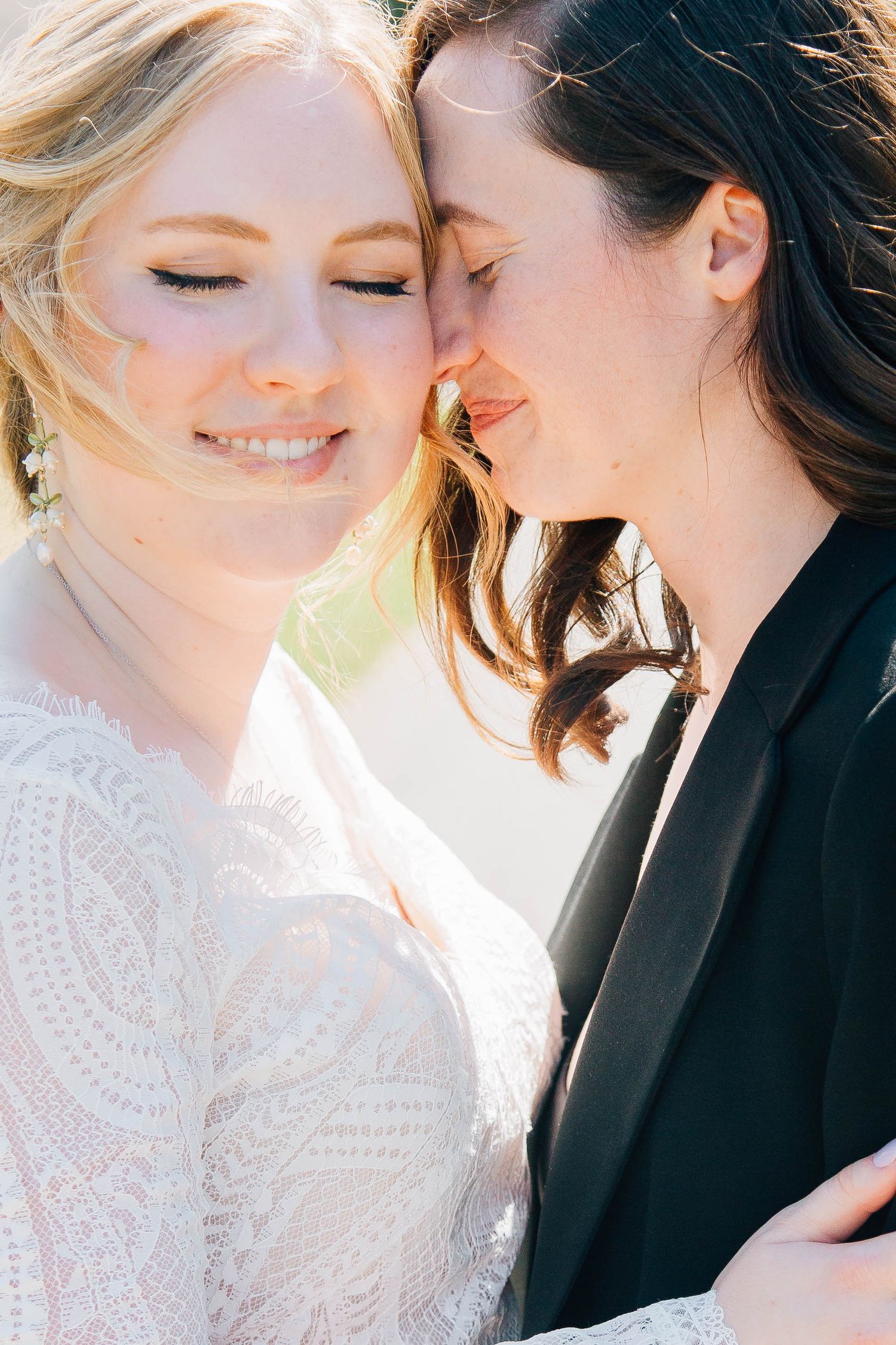 Two brides share an intimate moment, one in white lace dress and one in black blazer, touching foreheads on their wedding day.