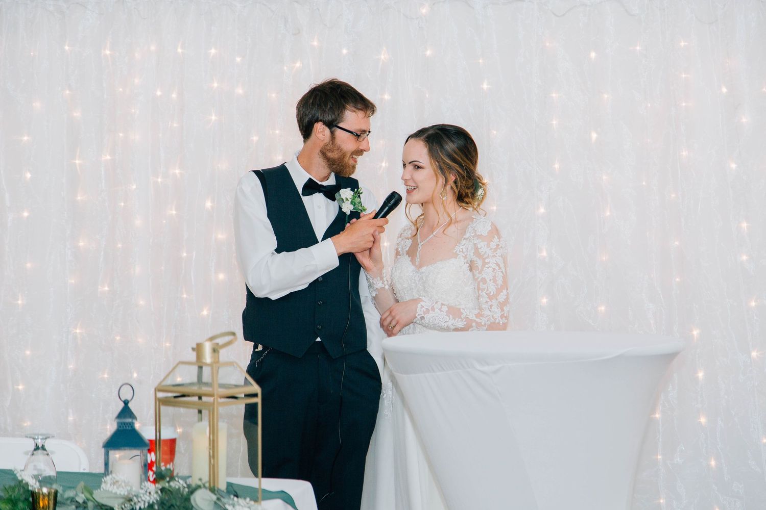 Smiling bride and groom share microphone while giving wedding reception speech at decorated venue.