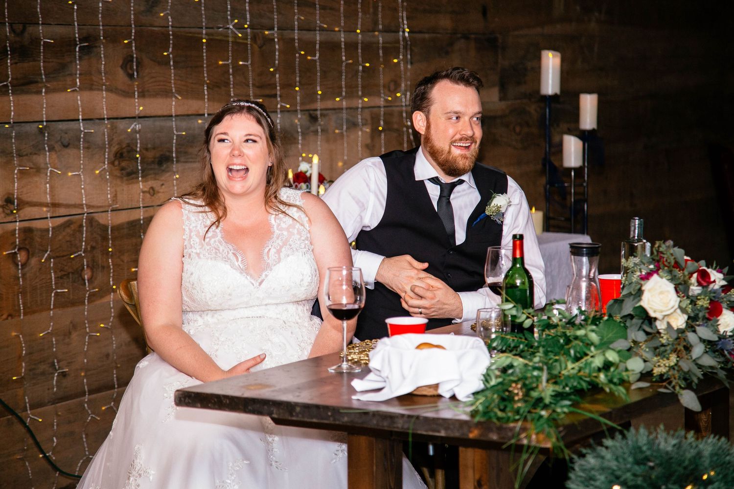 Bride and groom reacting with surprise and laughter at rustic barn wedding reception sweetheart table.