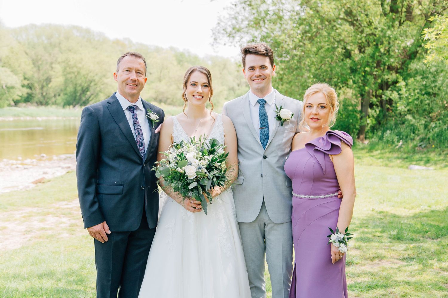 Bride and groom pose with parents near riverside, mother in purple dress and father in navy suit.