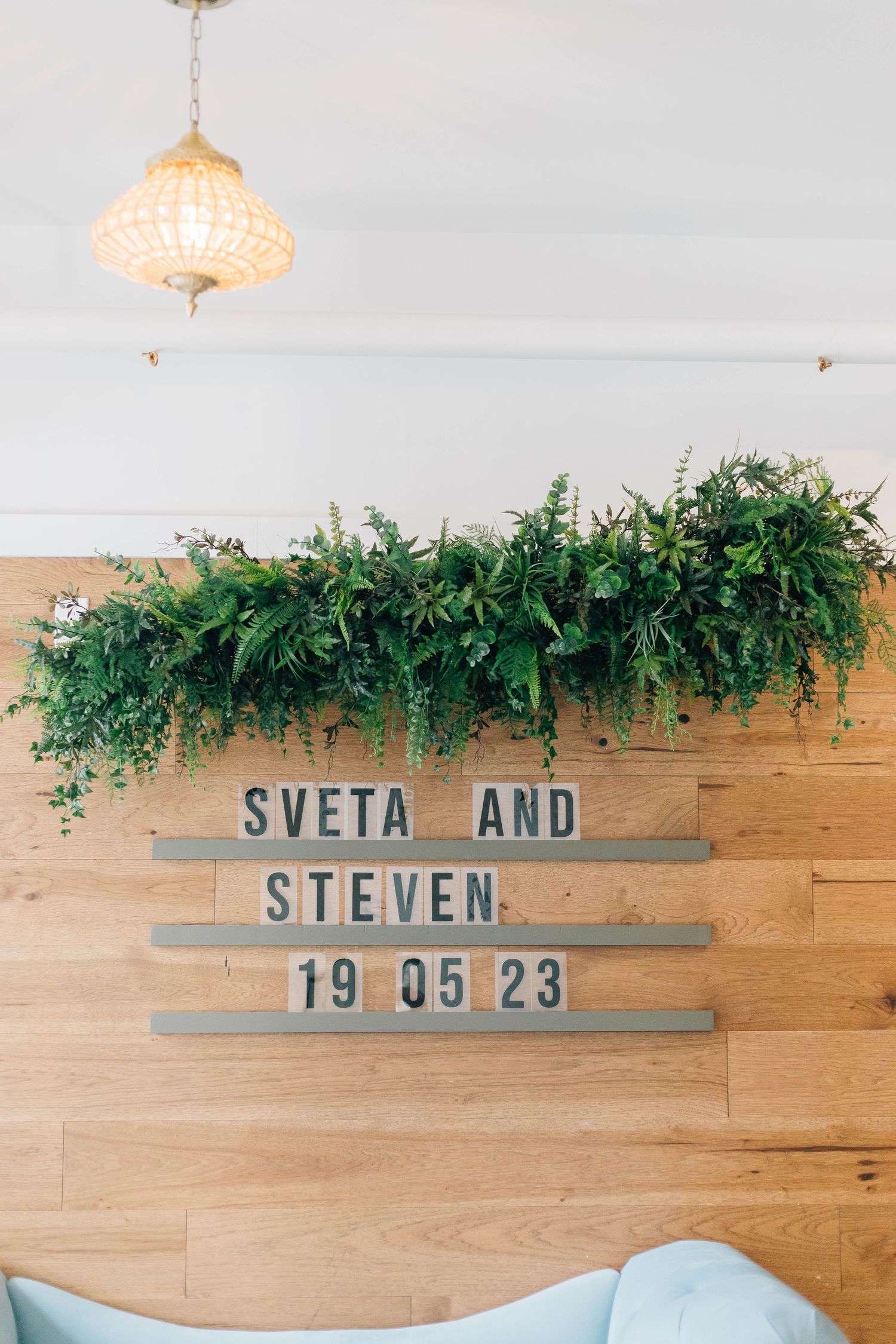 Wooden sign reading Sveta and Steven 19 05 23 decorated with lush green garland above.