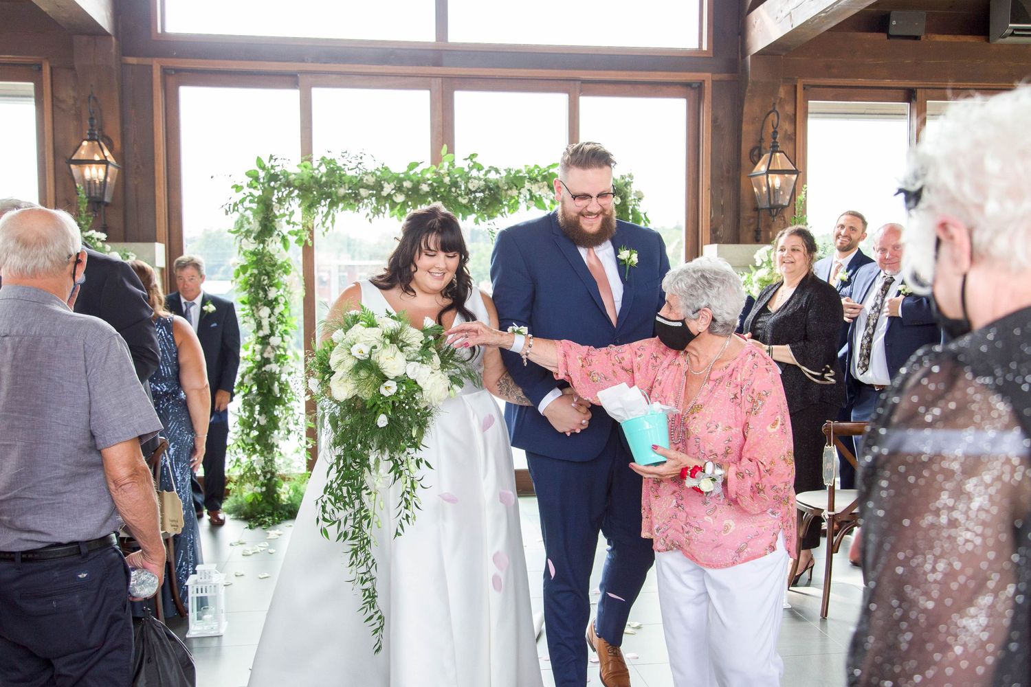 A newlywed couple walks down the aisle greeted by a guest in a rustic wooden ceremony venue.