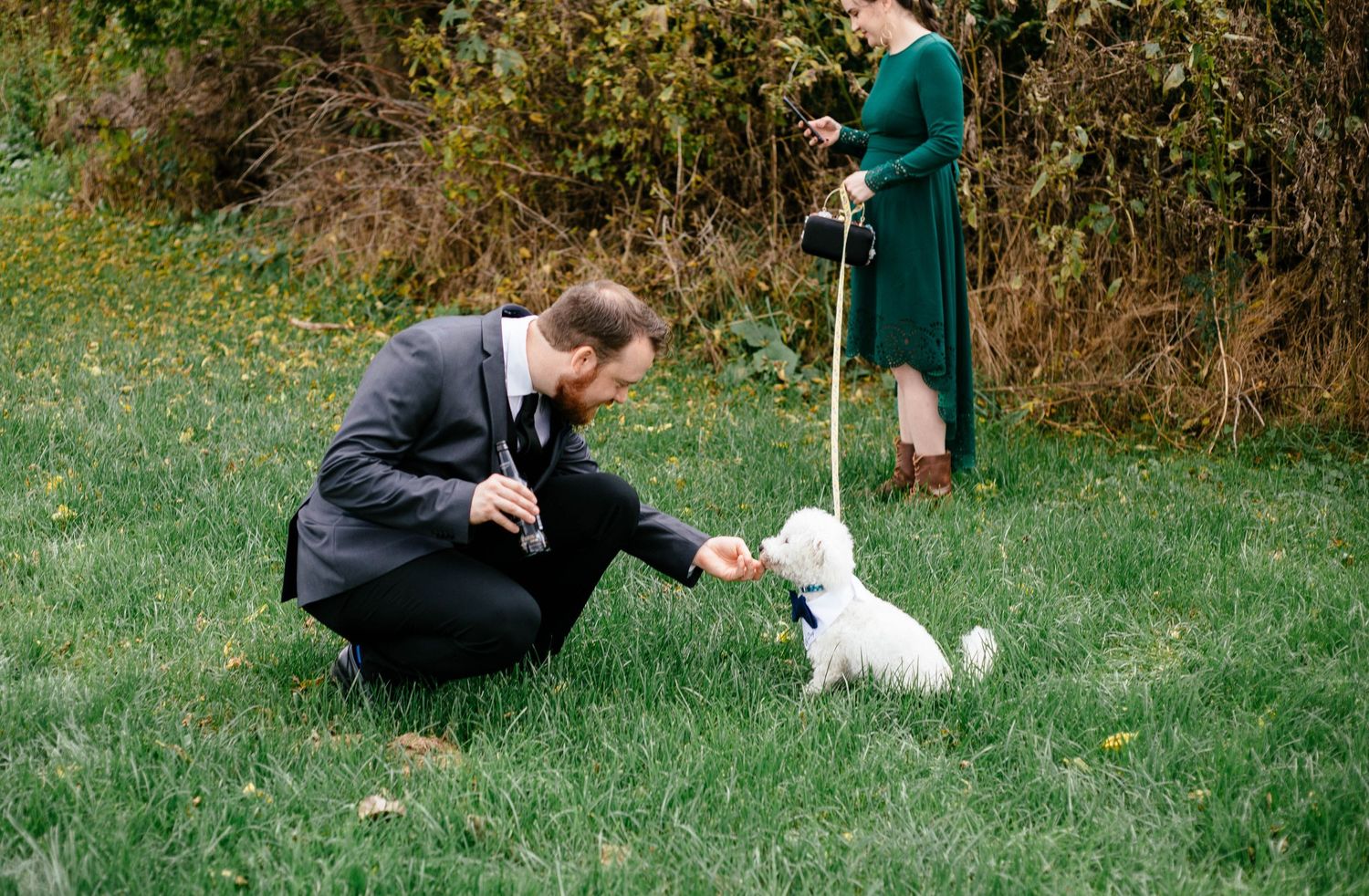 Groom in gray suit crouches to greet small white dog on leash held by woman in green dress outdoors.