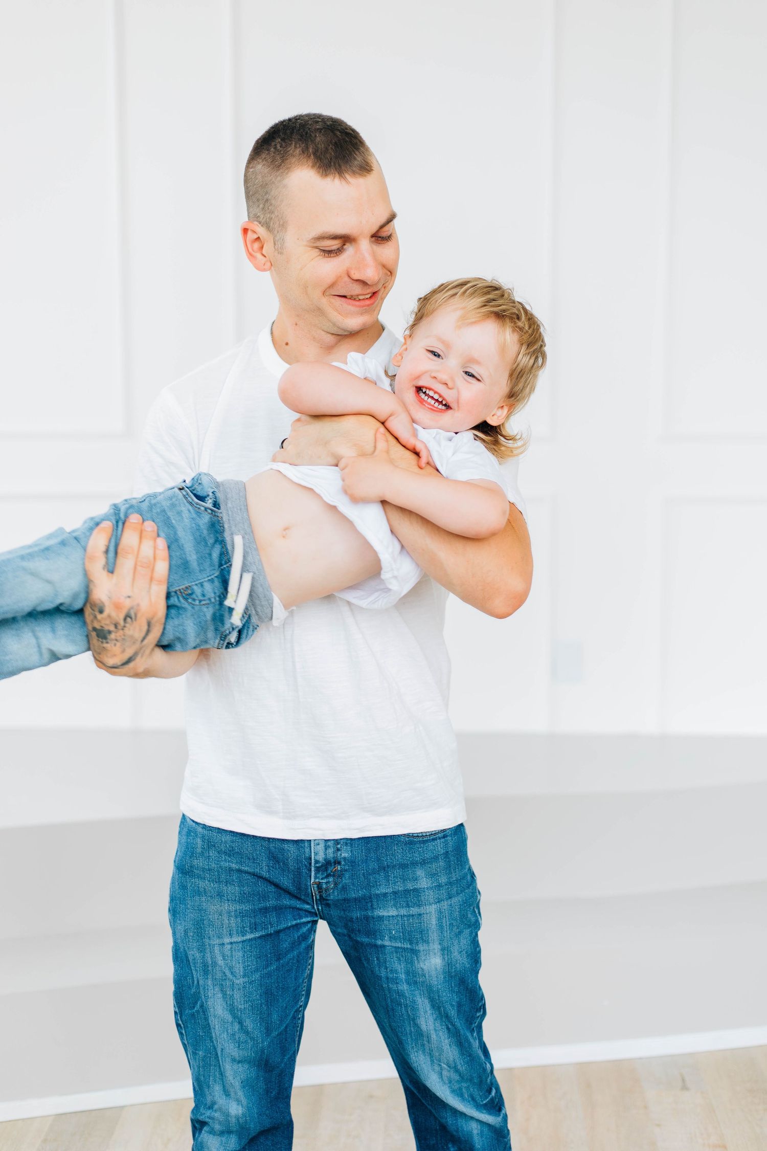Laughing tattooed father in white t-shirt playfully lifting giggling toddler son in bright studio.
