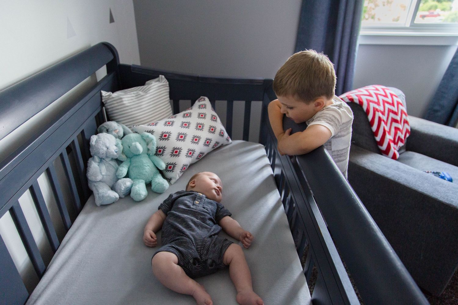 Young boy lovingly gazes at his newborn sibling lying in a navy crib.