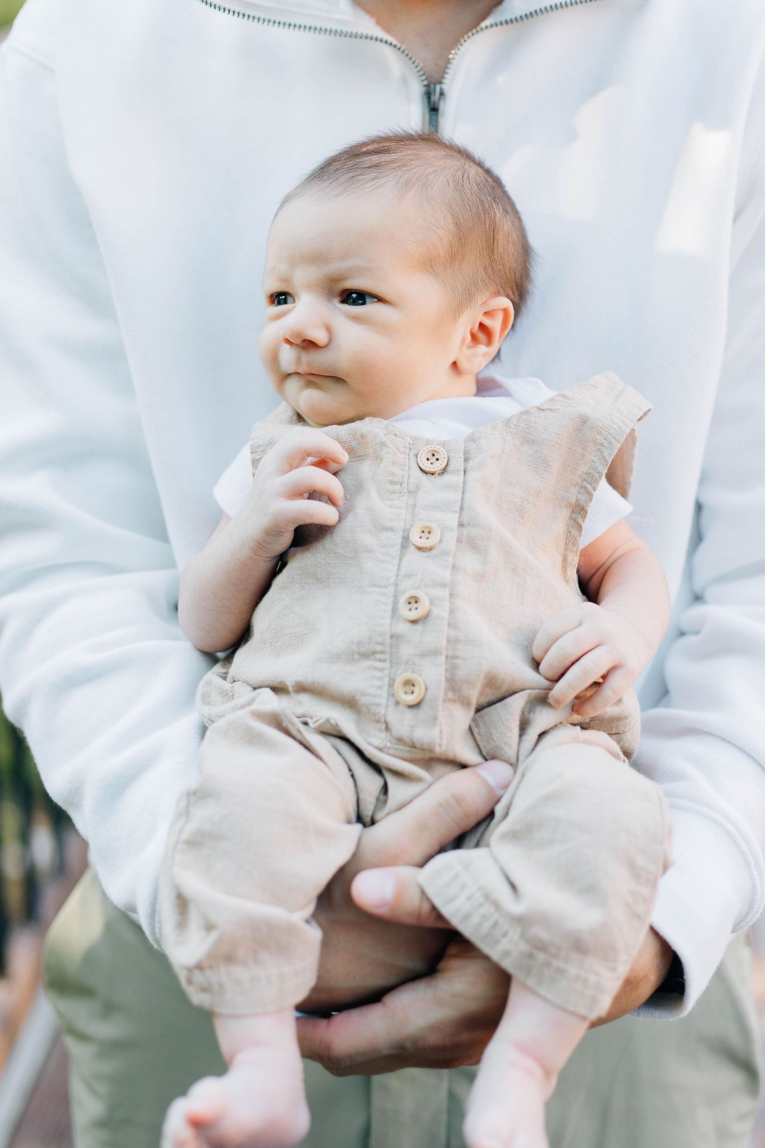 Father in white hoodie holding newborn baby dressed in beige linen romper outdoors.