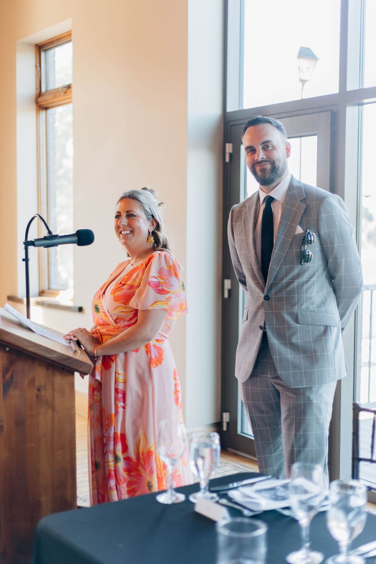 A woman in a floral dress speaks at a podium beside a man in a plaid suit at a wedding reception.