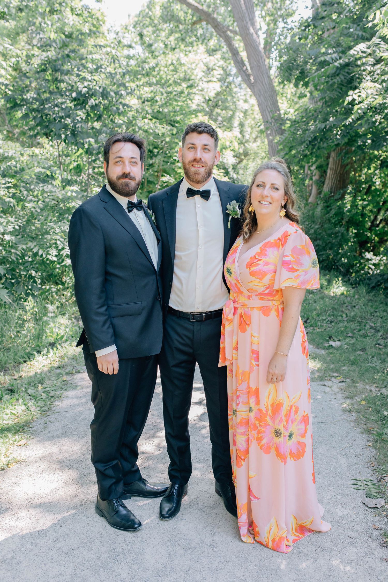 Groom in a black tuxedo stands with two smiling guests in formal attire on a tree-lined outdoor path.