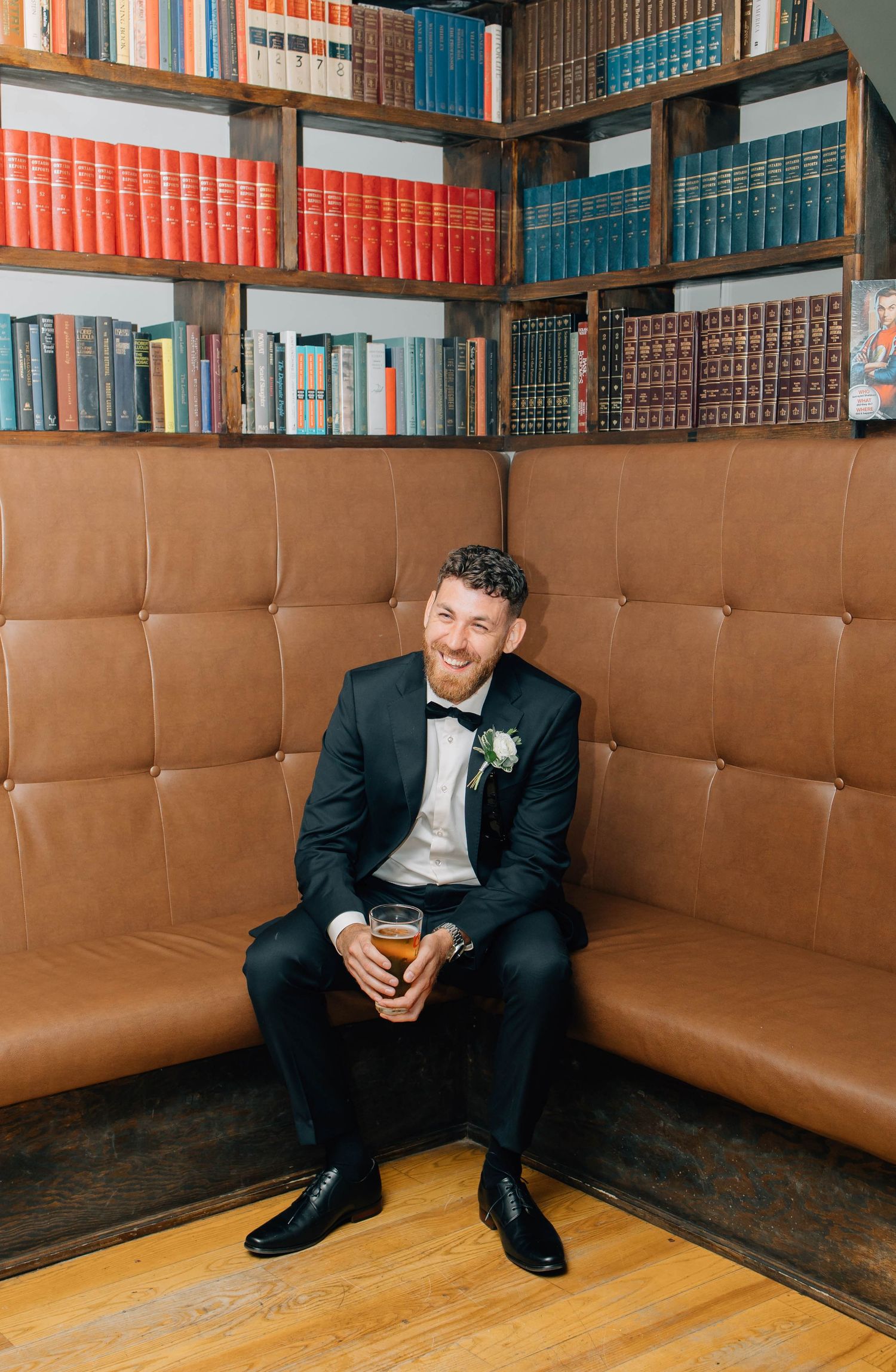 Smiling groom in black tuxedo sits on leather sofa holding a drink in front of bookshelves.