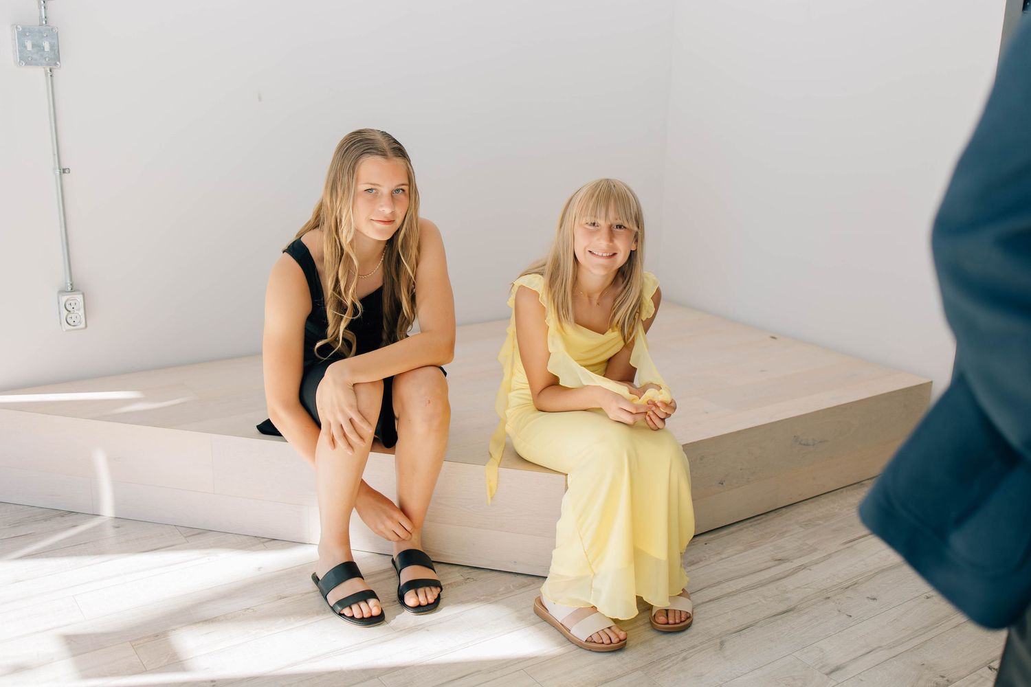 Two young female wedding guests in black and yellow dresses sit together on white platform indoors.
