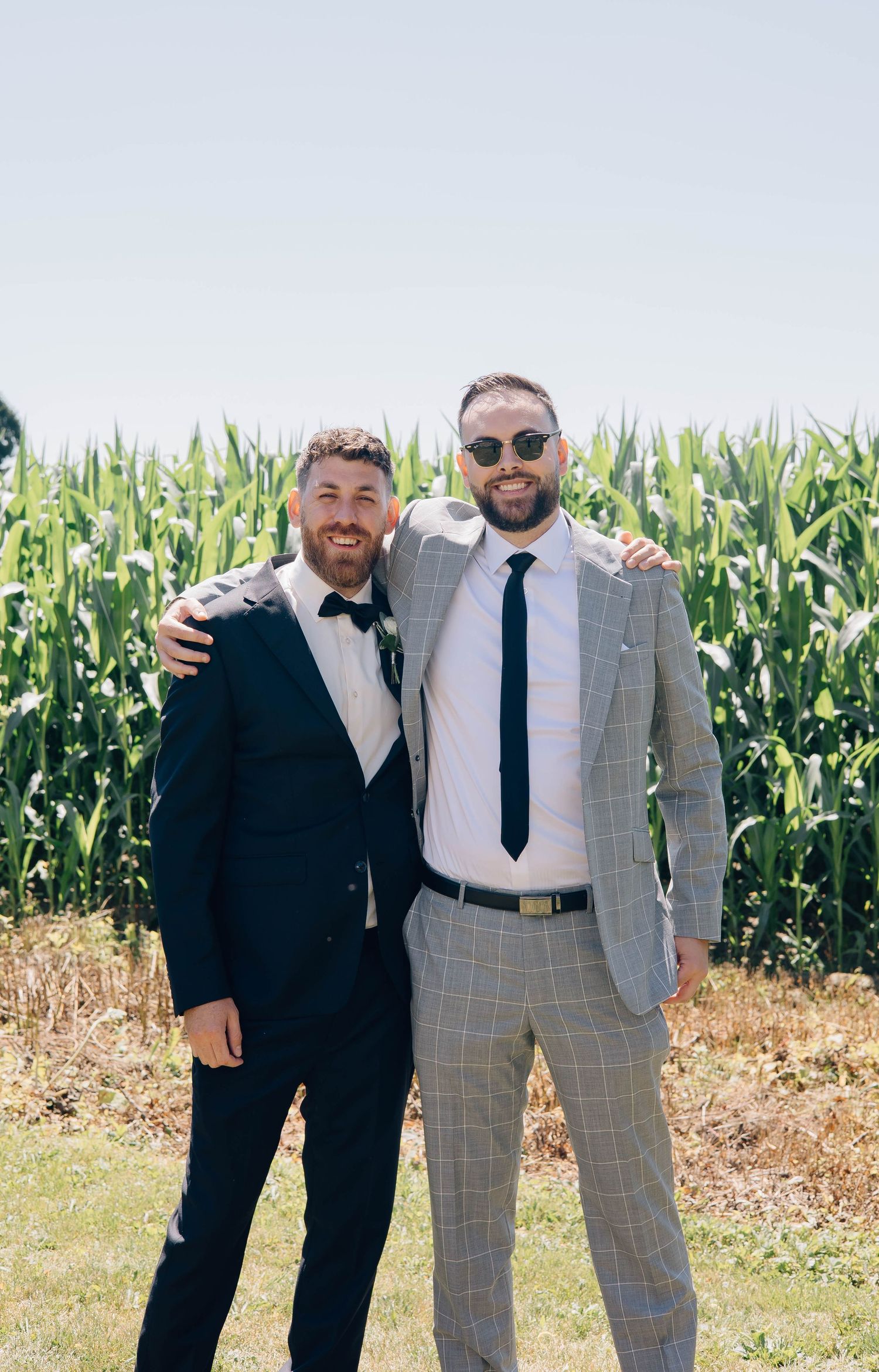 Two men in suits smiling with arms around each other in front of a green cornfield on a sunny day.