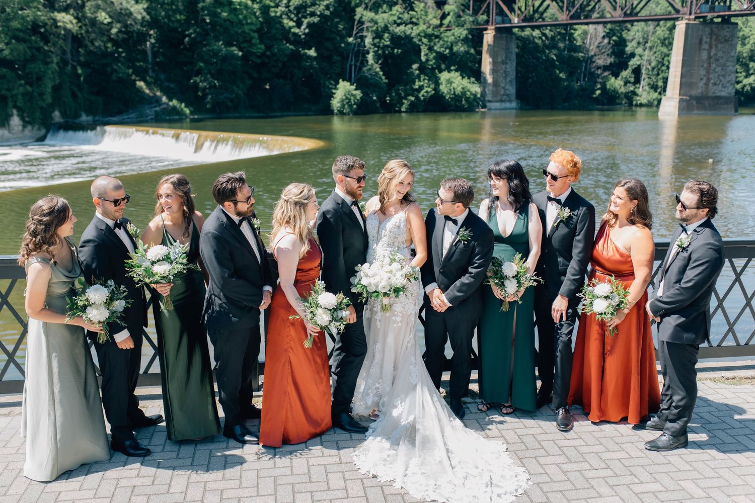 Wedding party posing together near a river bridge, bridesmaids in rust and sage gowns.