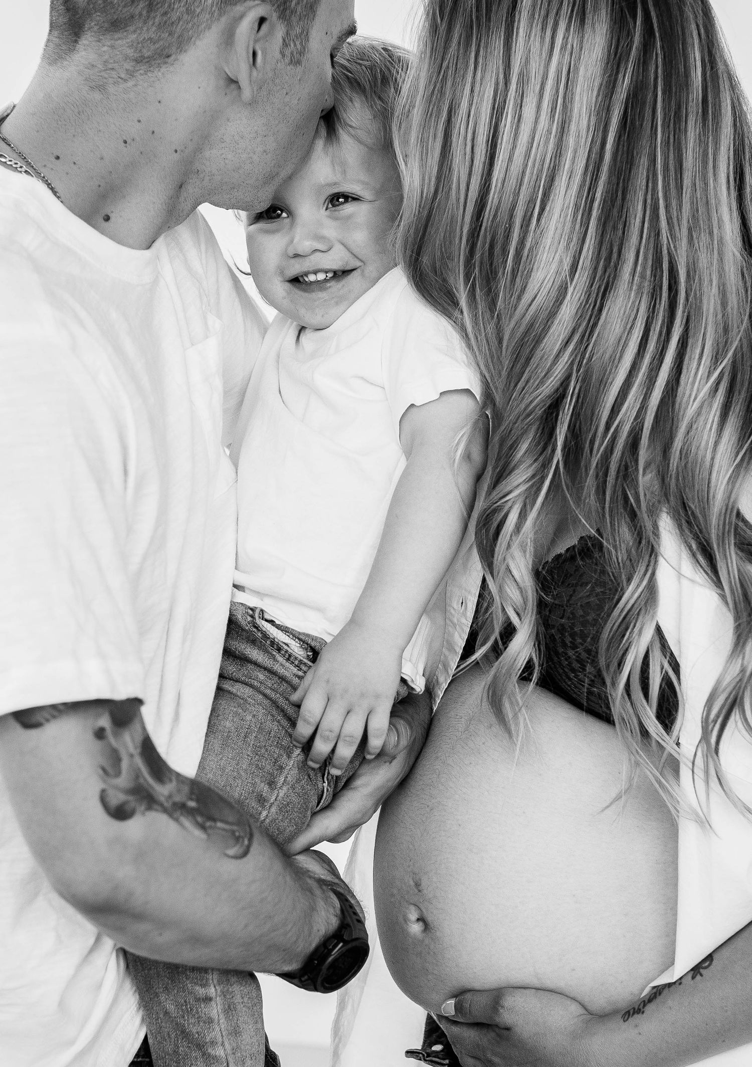 Black and white maternity photo of smiling toddler between parents with pregnant mom showing baby bump.