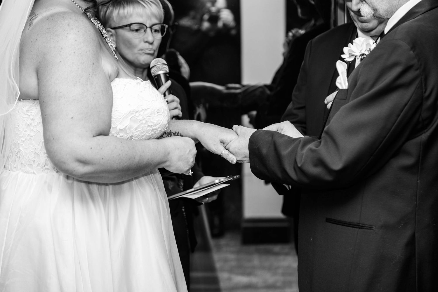 Black and white photo of bride reading vows while officiant holds microphone during wedding ring exchange.