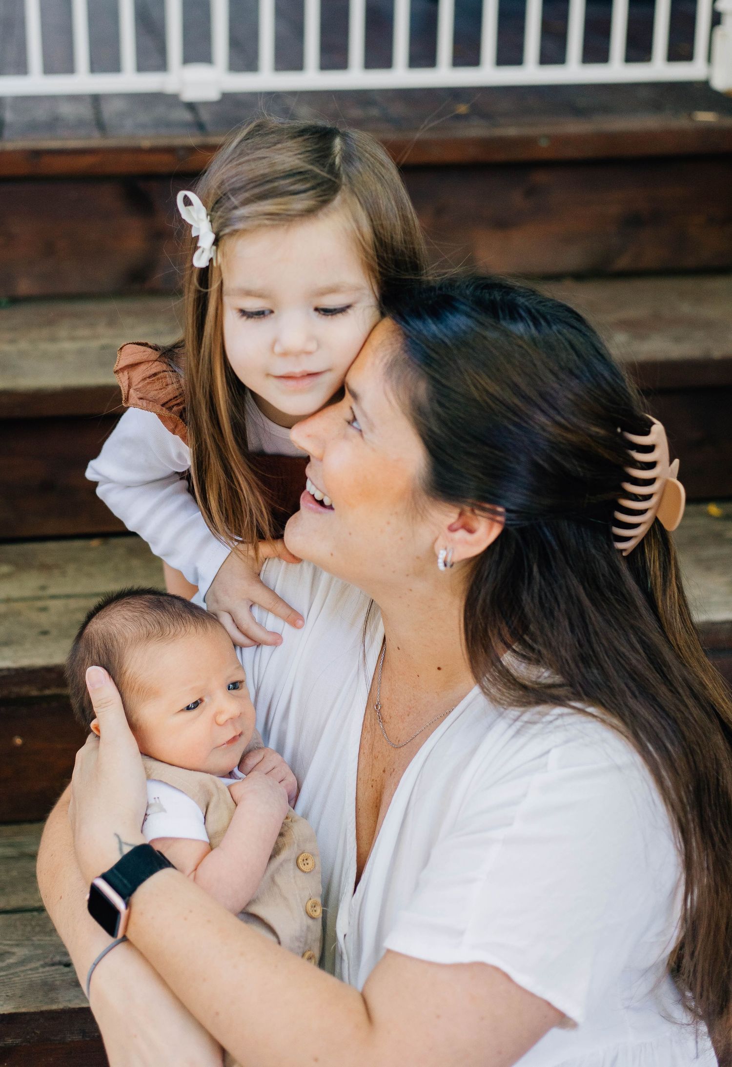 Young girl with white bow hugging her smiling dark-haired mother who cradles a newborn baby on porch steps.
