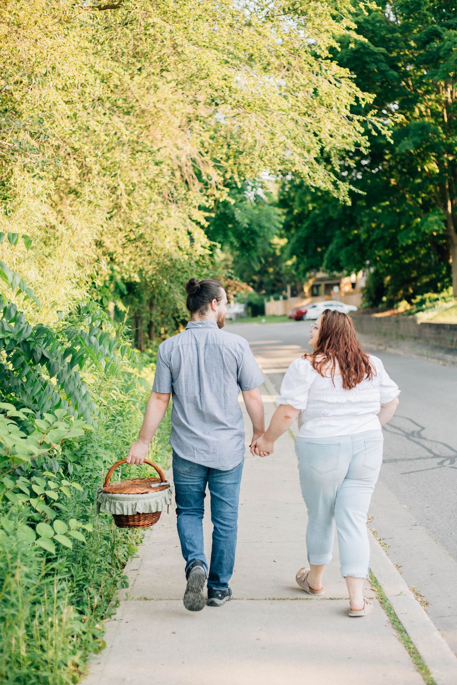 Couple walking hand-in-hand on a tree-lined sidewalk carrying a wicker picnic basket.