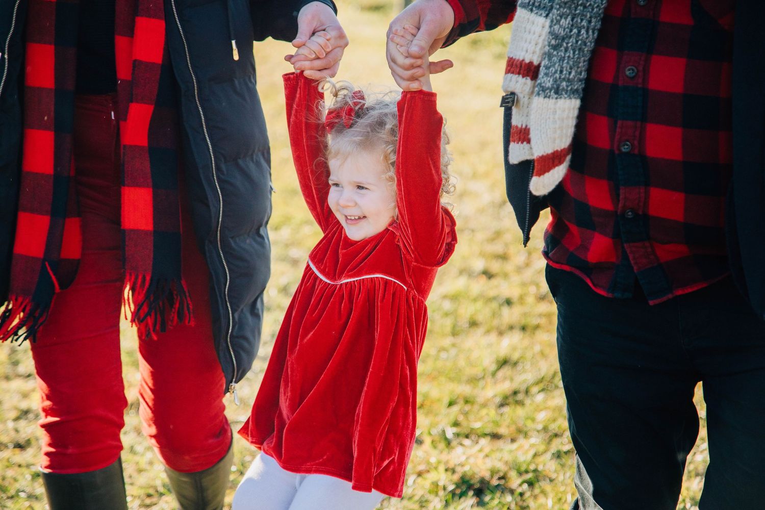 Toddler girl in red velvet dress held up by hands of parents wearing holiday outfits outdoors.