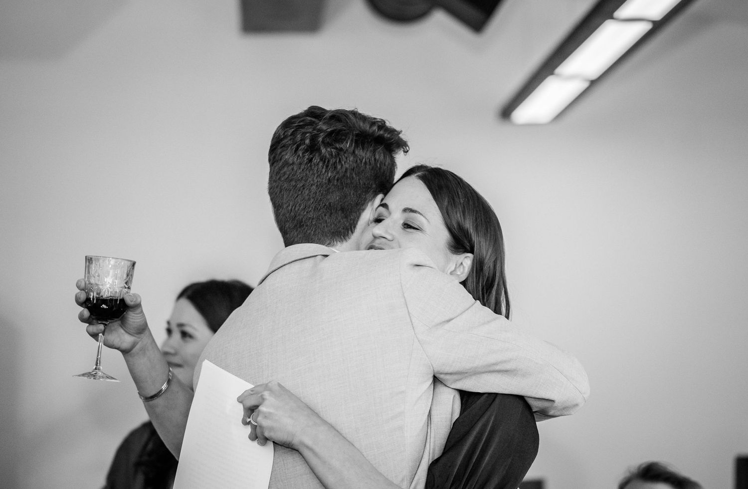 Emotional wedding guest hugging someone while holding a glass of red wine in black and white photo.