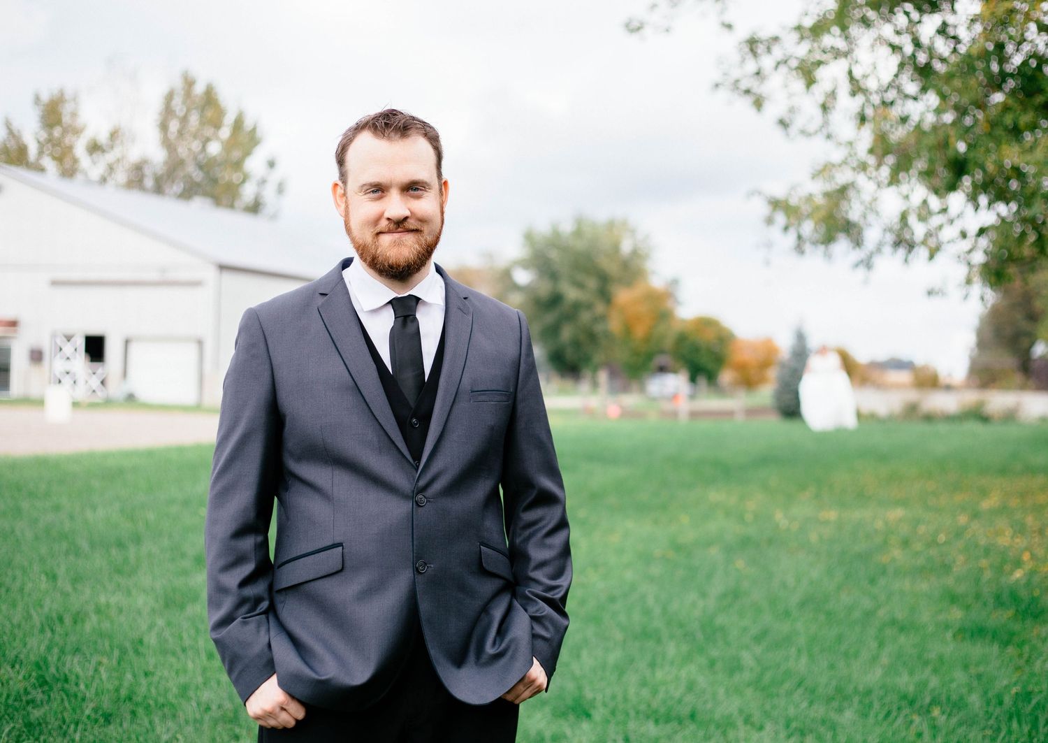 Handsome groom in gray suit and black tie standing on green lawn at wedding venue.