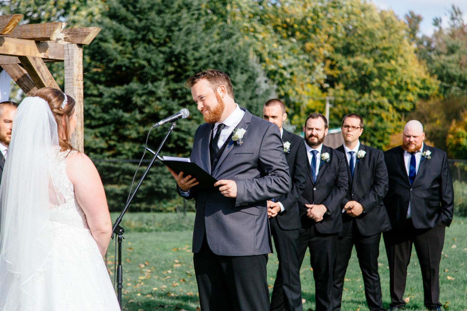 Groom reads vows at outdoor wedding ceremony with groomsmen standing behind him in navy suits.