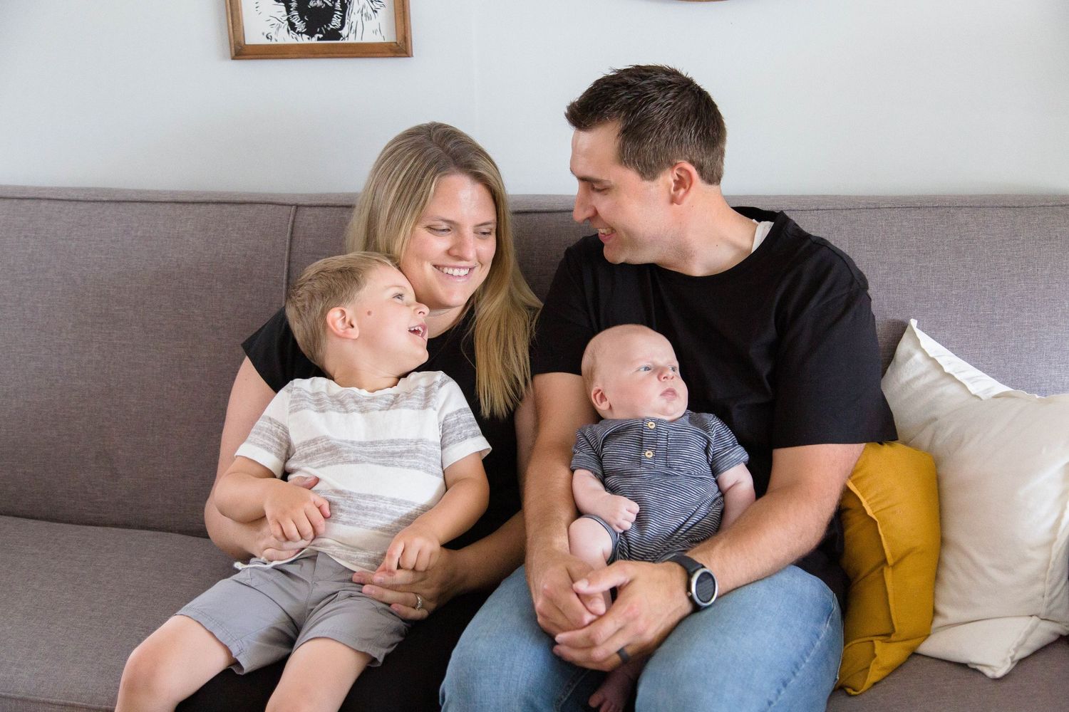 Happy family of four sitting on gray sofa, parents smiling at toddler son while dad holds newborn baby.