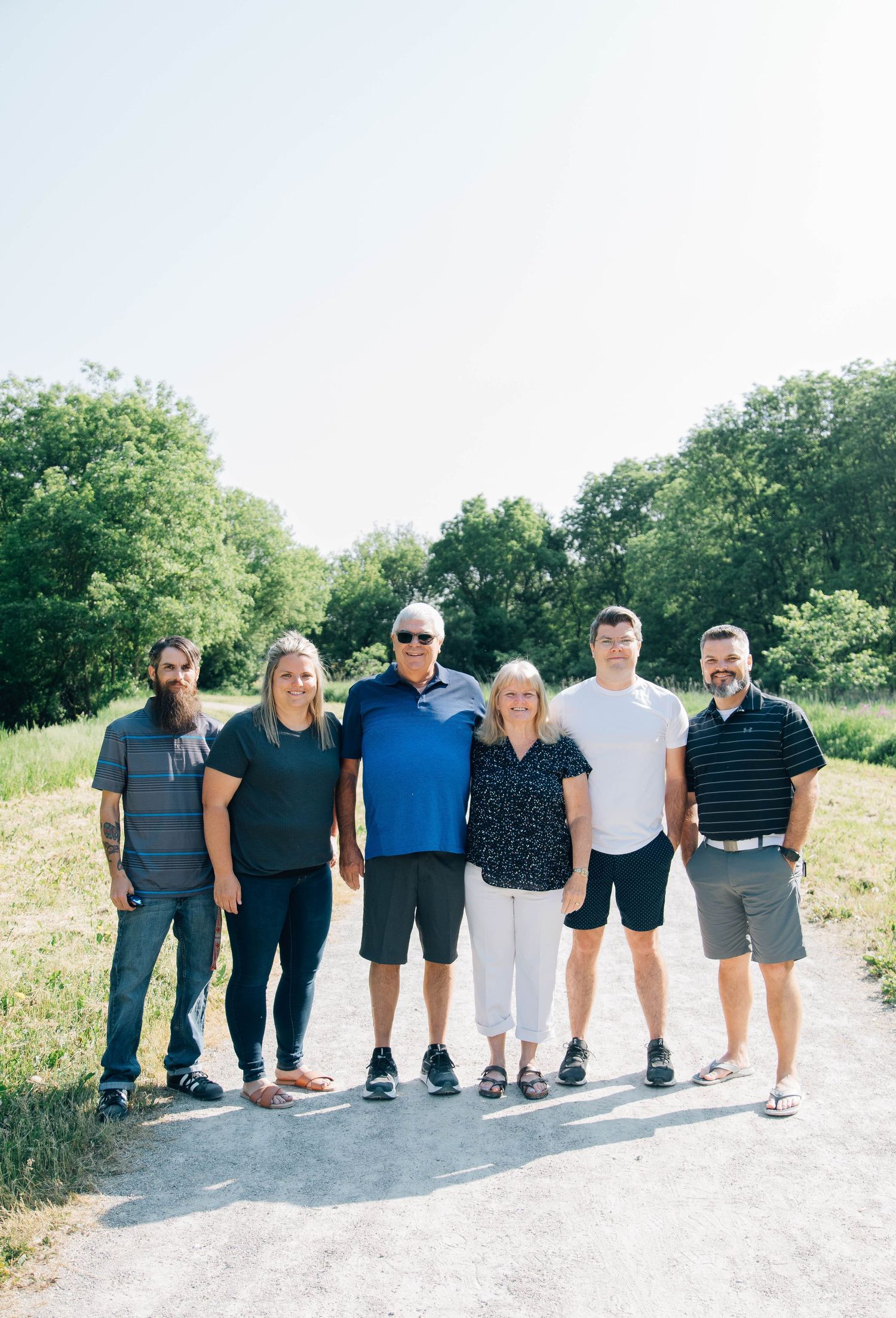 Group of six adults smiling together on a sunny outdoor path surrounded by green trees and open sky.