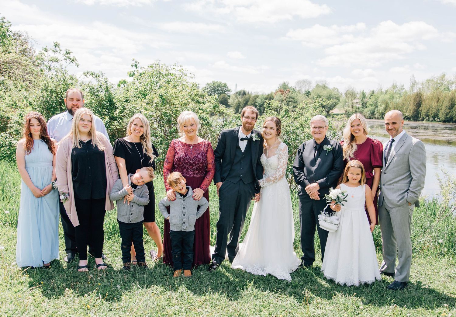 Large wedding family portrait group standing on green grass beside a peaceful pond on a sunny day.