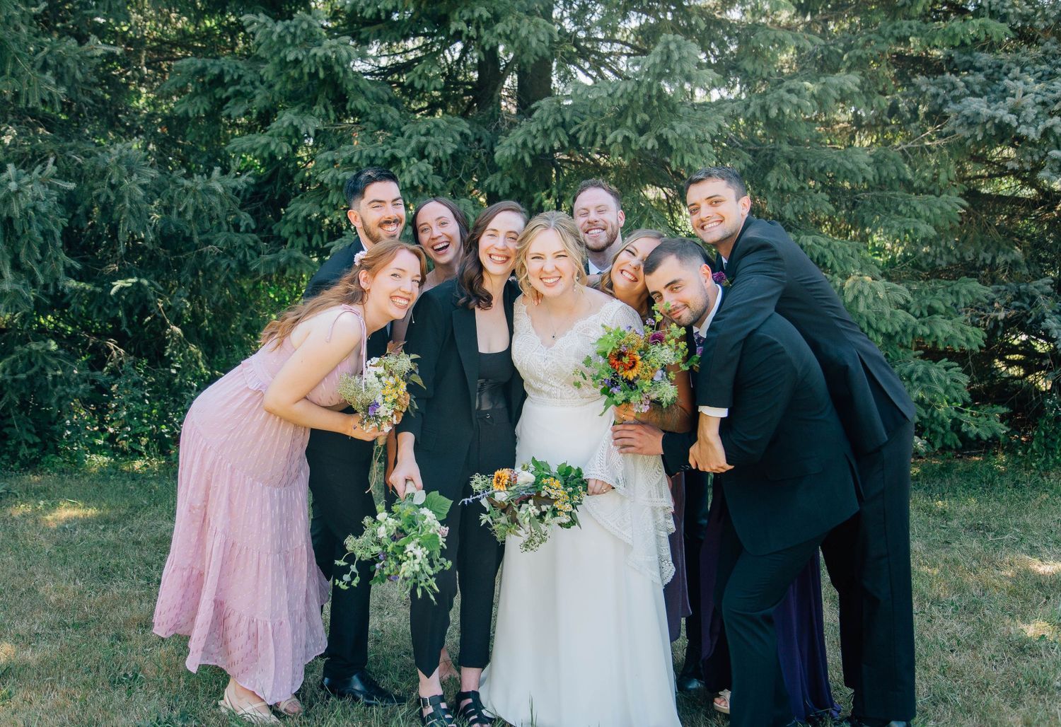 Bride surrounded by smiling wedding party members huddled together in a joyful group pose outdoors.