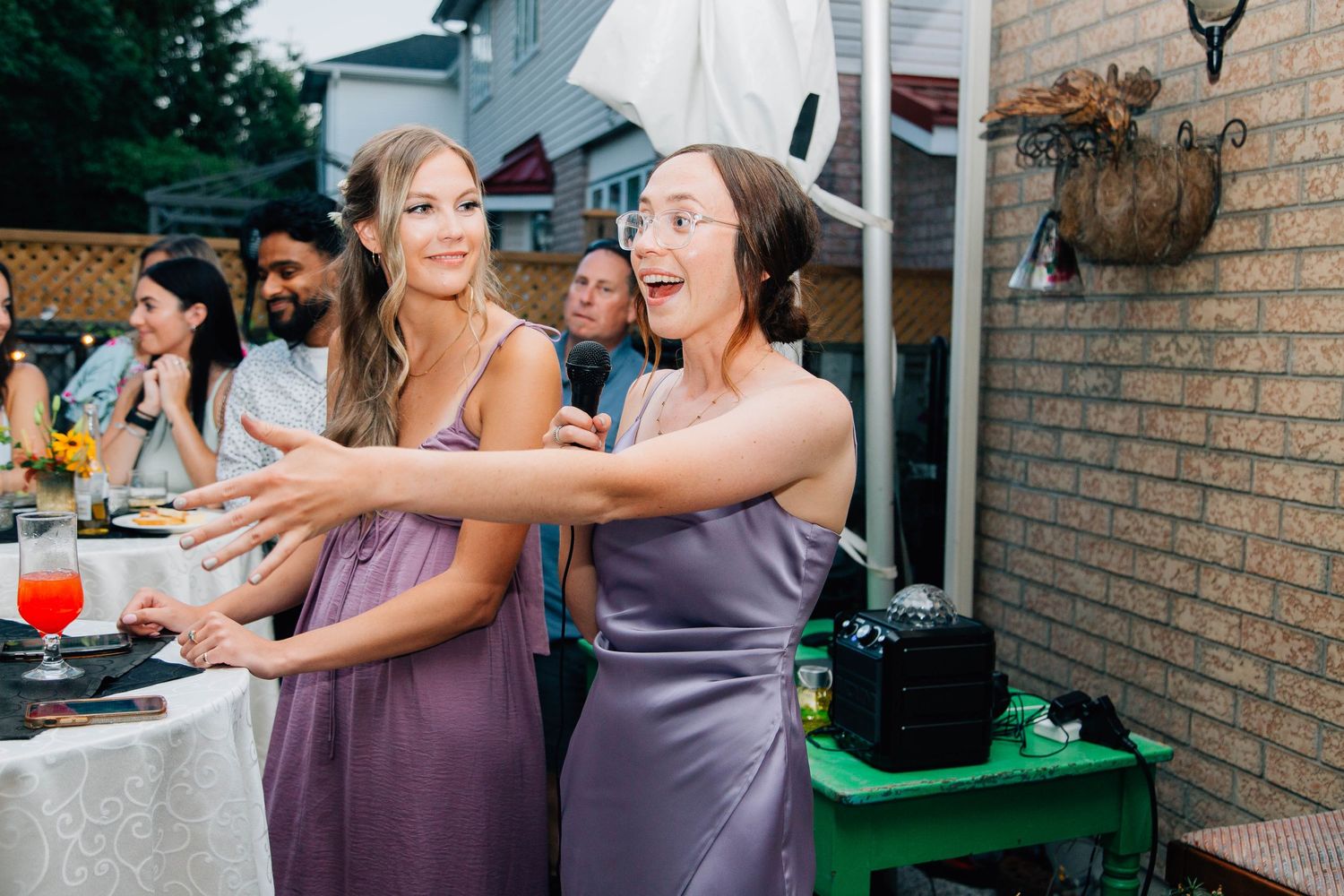 Two bridesmaids in purple dresses laugh and dance together at an outdoor wedding reception.
