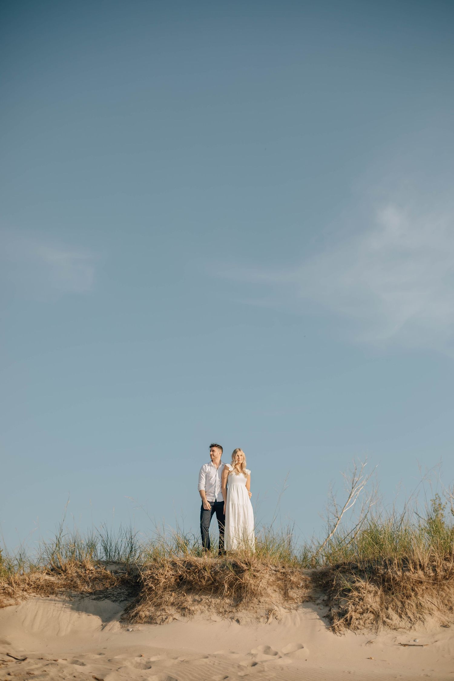 Couple standing on sandy dunes under blue sky, woman in white dress and man in casual attire, romantic outdoor portrait.