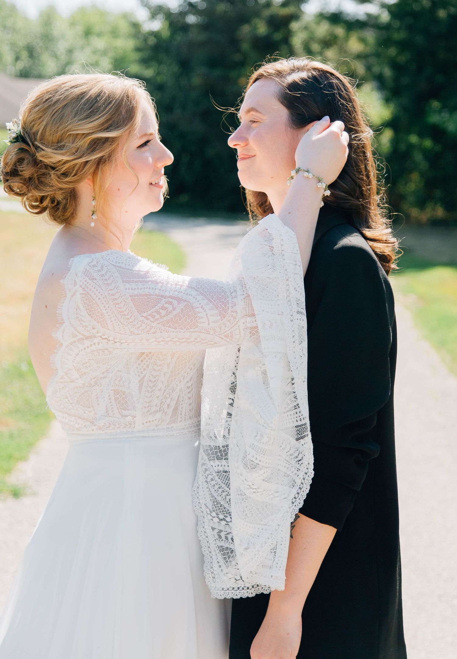 Bride in lace gown gently touches companion's face as they share an intimate moment on a sunny path.