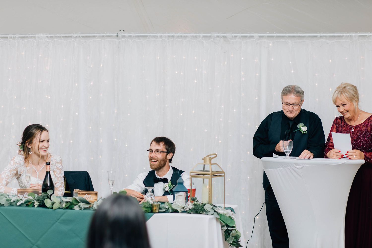 Two family members give joint toast at wedding reception as happy couple listens at head table.