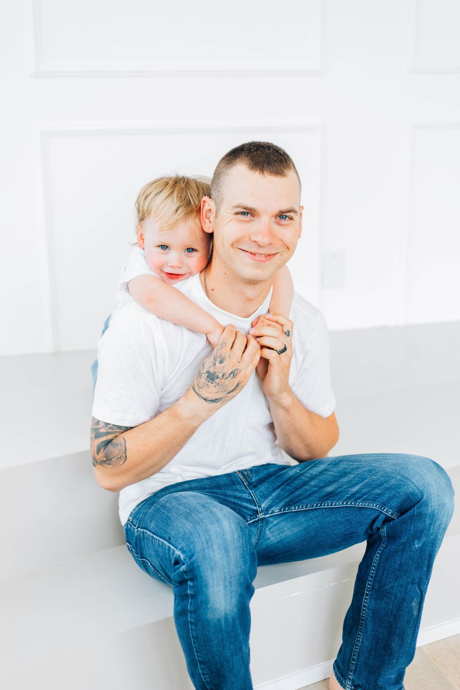Smiling tattooed father in white tee and jeans with toddler hugging him from behind on bench.