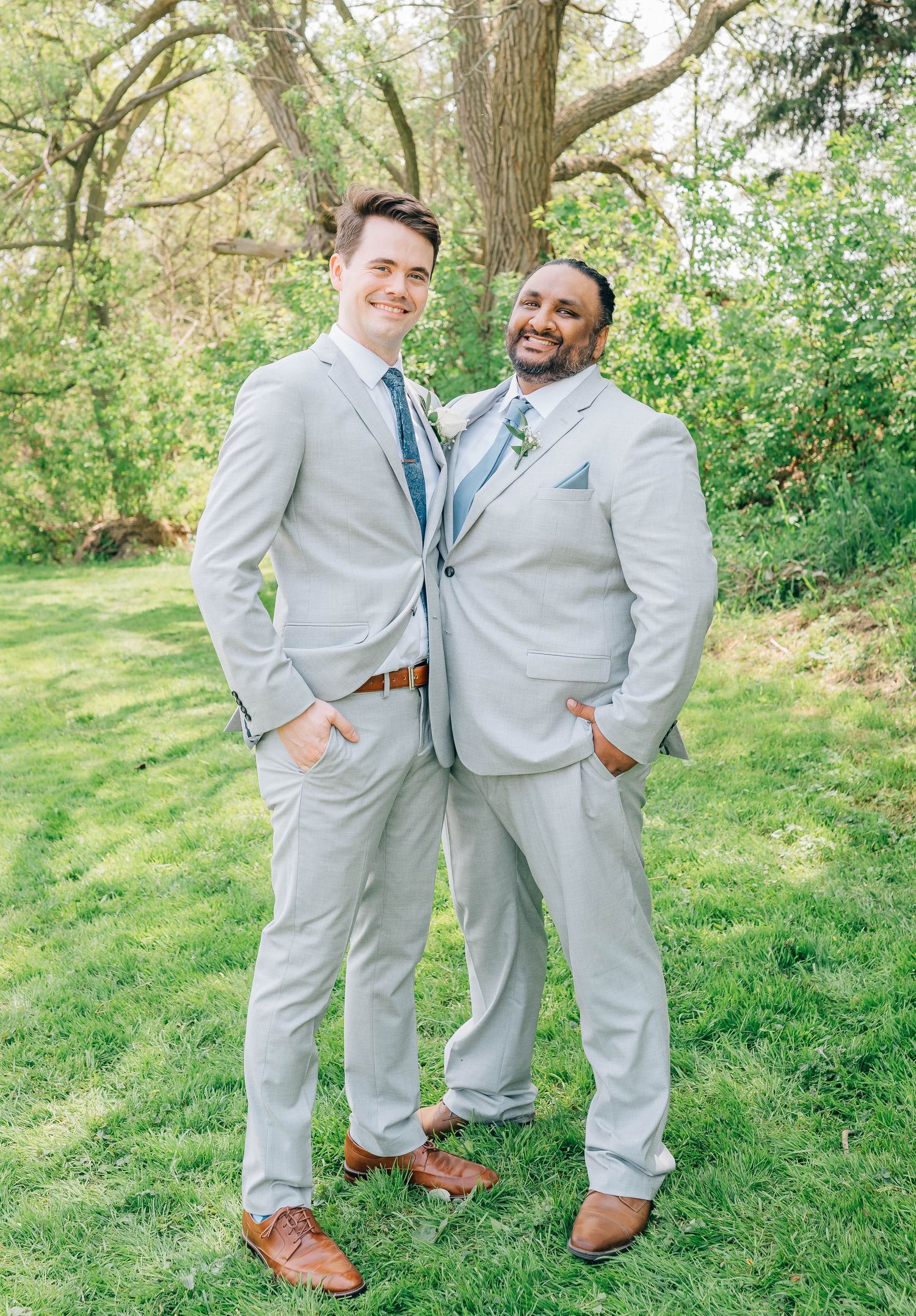 Two groomsmen in light gray suits smile and pose together on green lawn beneath large tree outdoors.