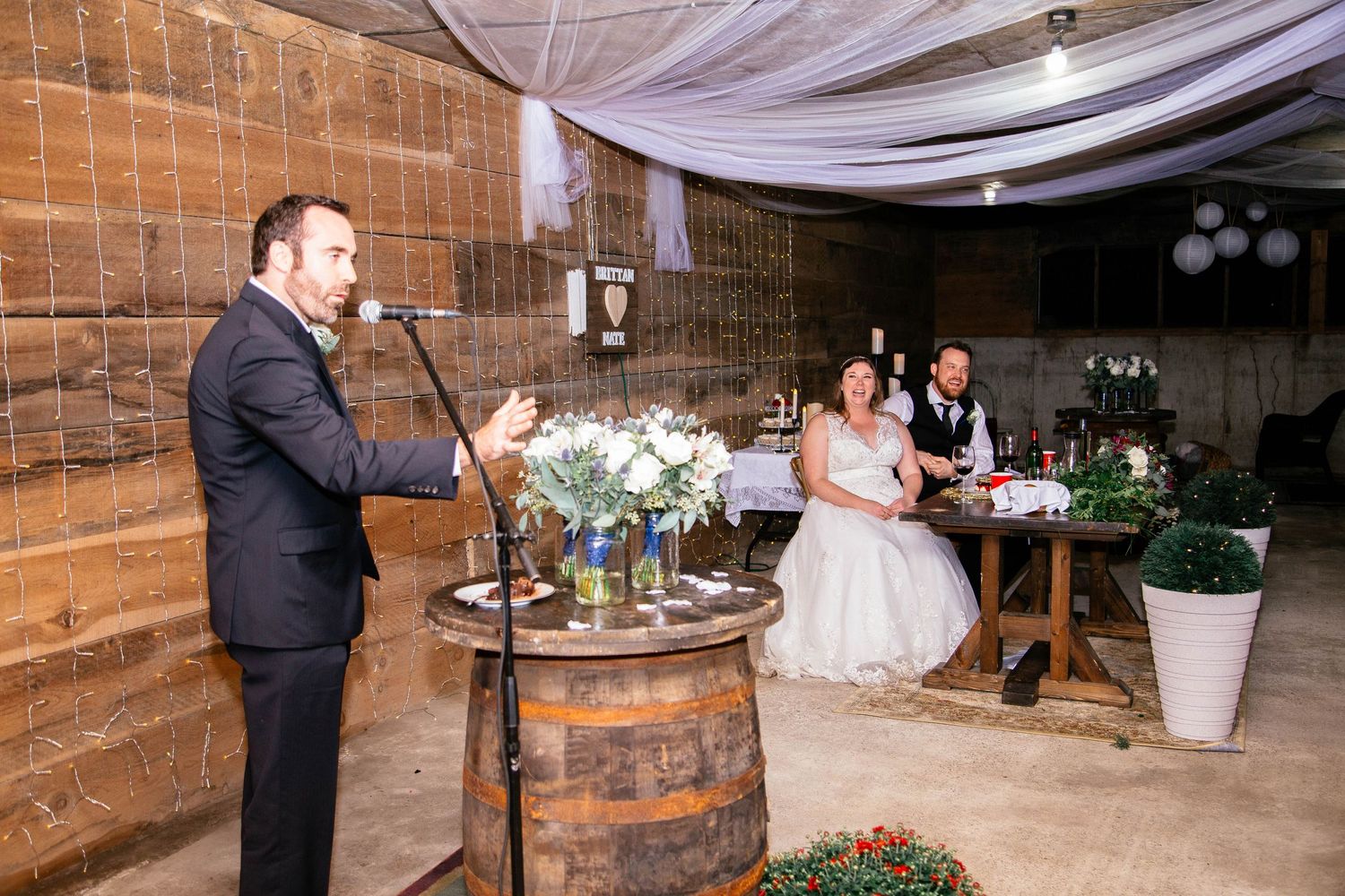 Best man giving toast at rustic barn wedding reception while bride and groom listen attentively.