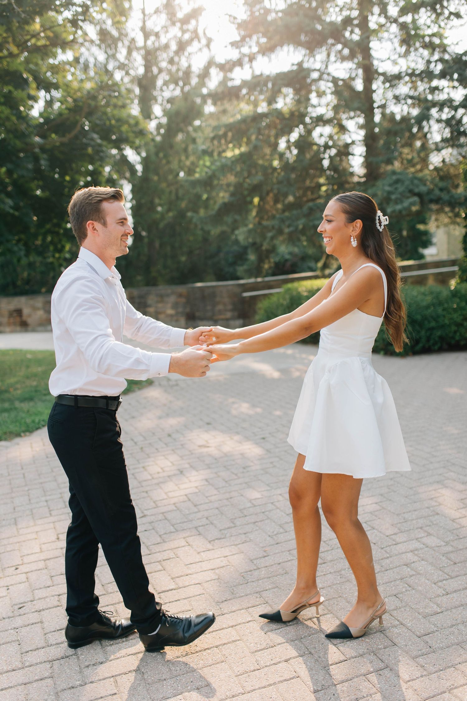 Engaged couple holding hands and dancing on brick path, woman in white mini dress and man in white shirt.