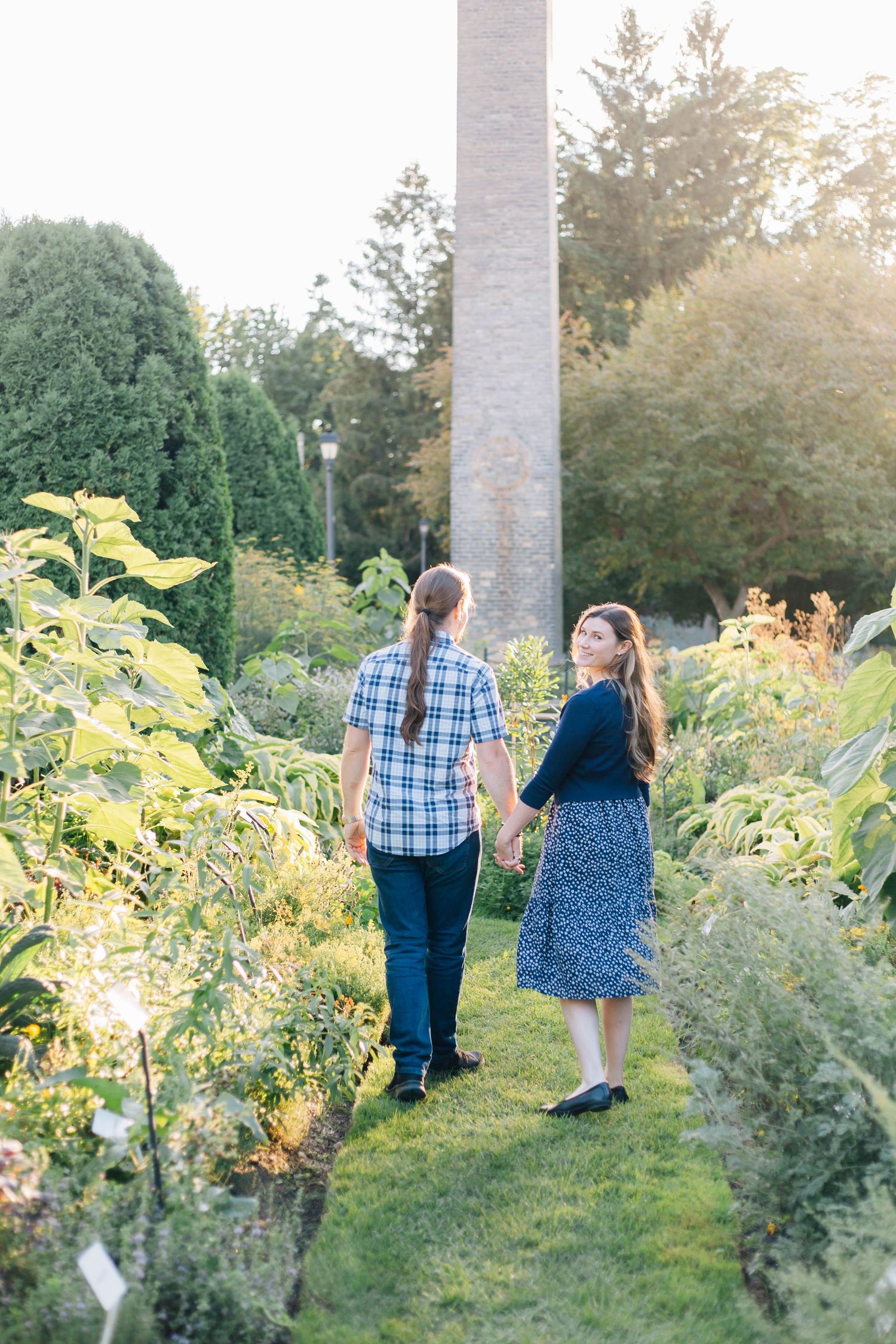 Couple walking hand in hand through sunlit botanical garden, man with ponytail in plaid shirt and woman in navy dress.